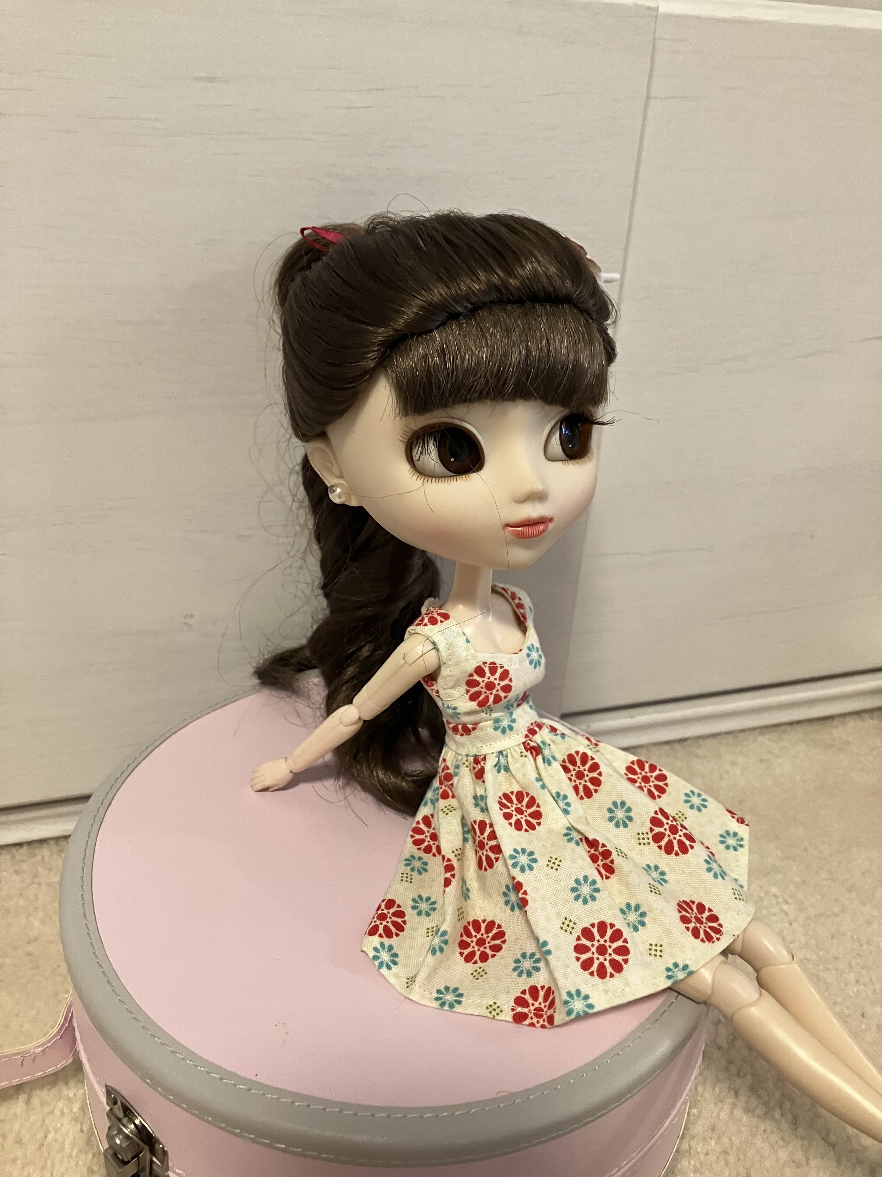 a photo of a pullip doll, which is a one-sixth scale collector doll with a very large head. this particular doll has brown hair with short bangs that is worn in a long, loosely curled ponytail. her eyes are also brown, and her skin is pale. she is looking to the side, away from the camera. she wears an off-white colored sweetheart dress printed with red, blue, and green flowers, and she sits atop a round pink box.