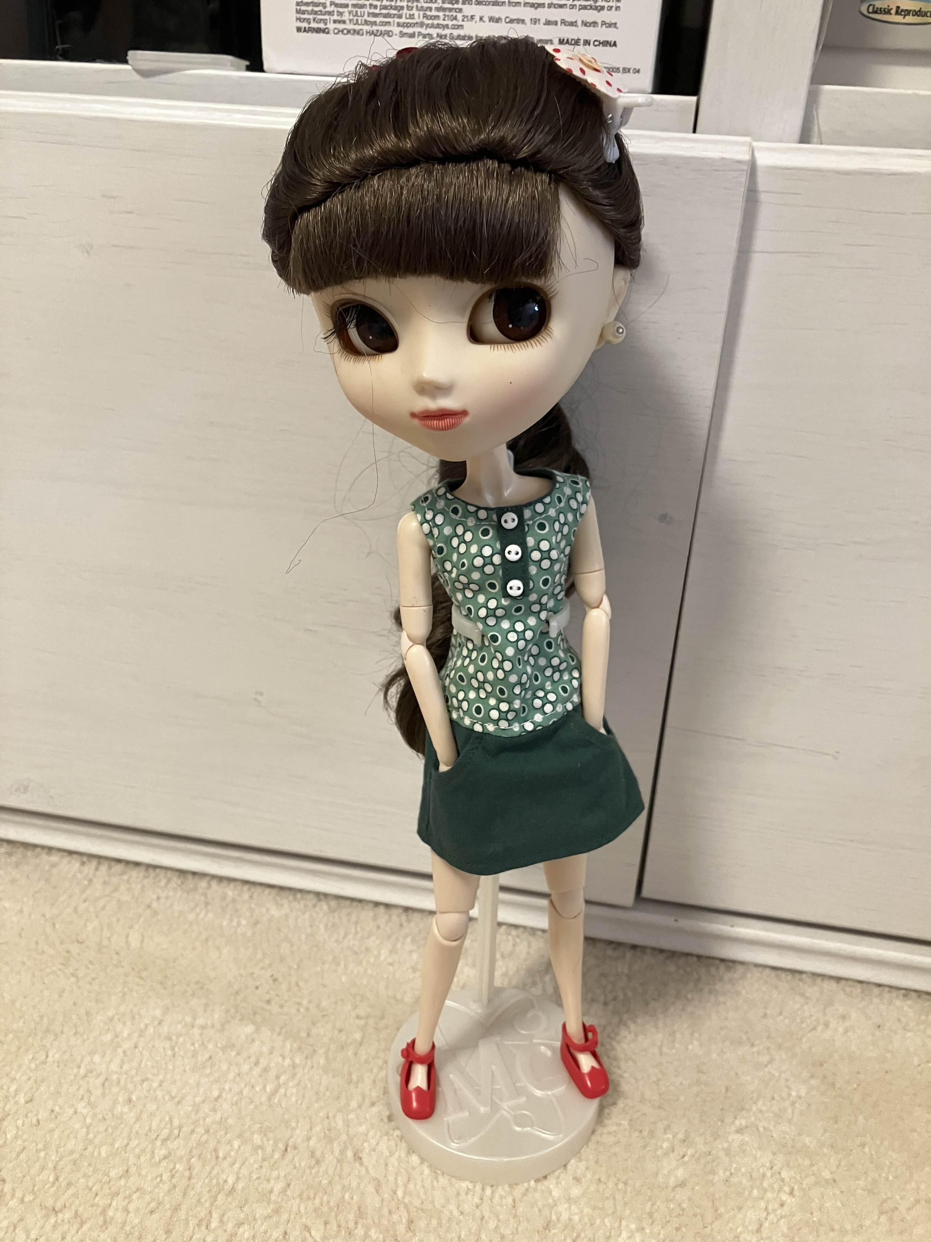 a photo of the same pullip doll, now standing with the help of an admittedly flimsy plastic stand. in this picture she wears a different dress; this one green with a white dotted pattern that stops at the waist, making the skirt solid green. the dress has tiny pockets; the doll stands with her hands in the pockets.
