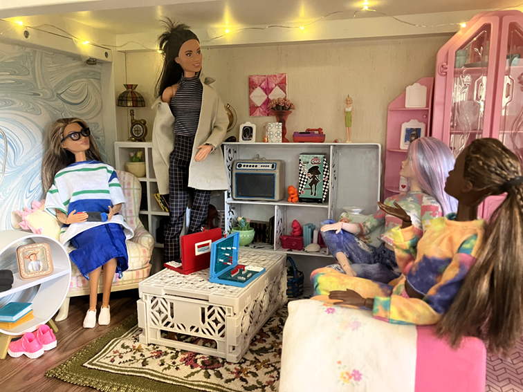 a picture of four barbie dolls in a living room-like diorama set up inside a bookcase. this alt text is under construction sorry
